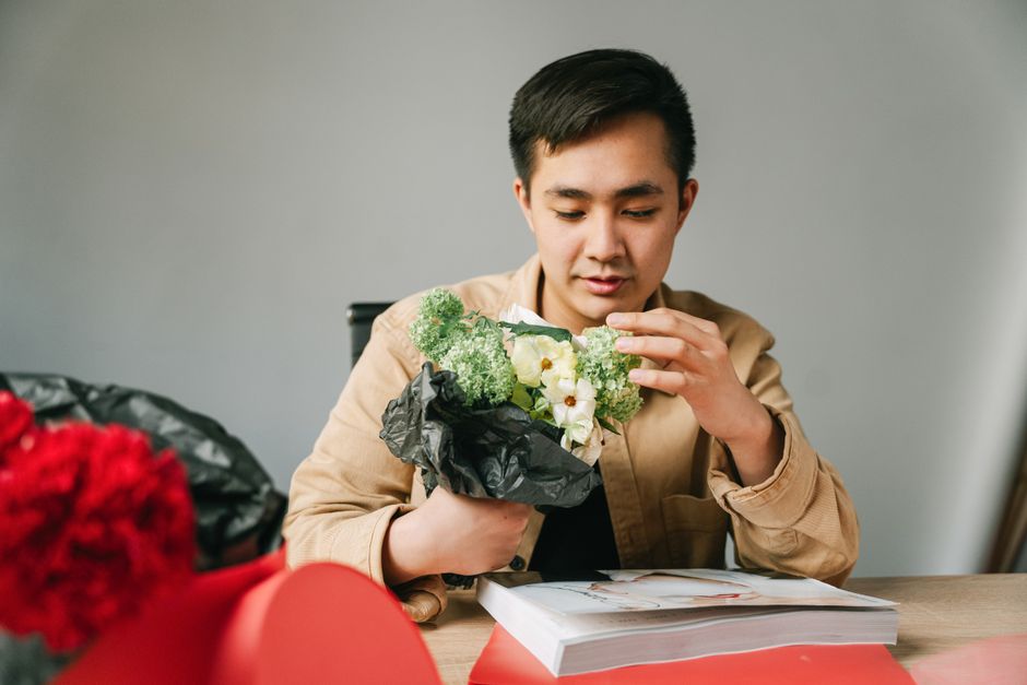 A young man with dark hair, wearing a beige jacket, is seated at a wooden table in a neutral-colored room. He is carefully examining a small bouquet of fresh flowers, which includes white blooms with 