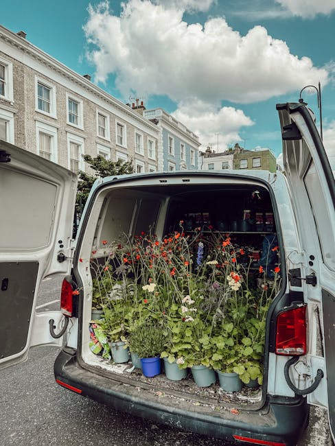 The open rear of a white van filled with a variety of flowering plants, including red poppies, white daisies, and green foliage, arranged in light blue pots. The flowers are fresh, with delicate petal