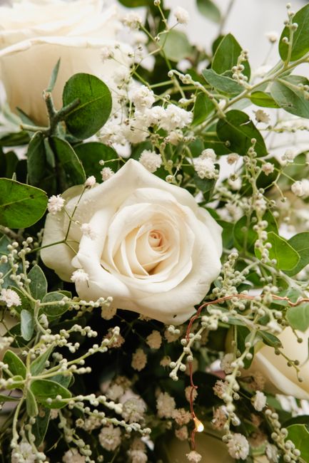 A close-up view of a wedding floral arrangement featuring a single white rose with delicate, softly-curved petals, surrounded by sprigs of small white gypsophila flowers and lush green eucalyptus leav