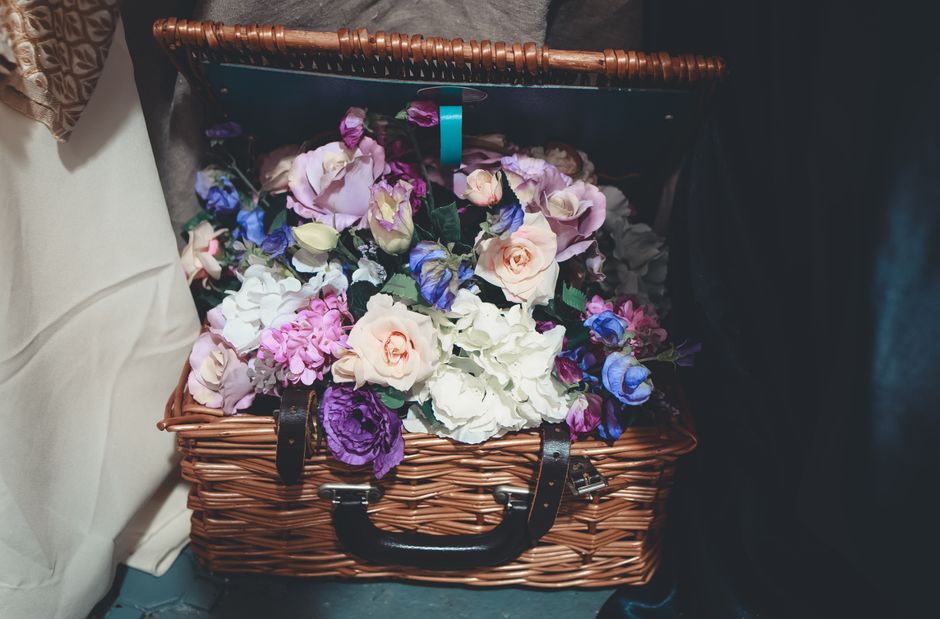 A woven wicker basket filled with a vibrant floral arrangement placed on the floor at an indoor setting. The bouquet includes large, soft pink roses, white hydrangeas, and purple and blue flowers, arr