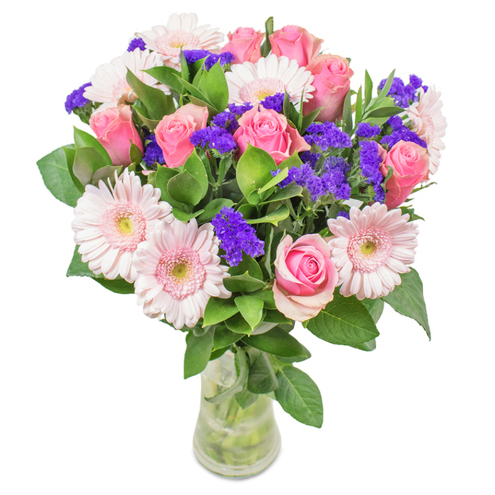 A vibrant bouquet, elegantly arranged in a clear glass vase and photographed against a crisp white backdrop, captures the energetic diversity of Brixton. The arrangement centres on an inviting cluster of seven to nine tightly furled and just-opening roses in delicate shades of pink, each bloom unfolding in an iconic spiral as if echoing the whorled wrought-iron designs on old Atlantic Road balconies. Complementing them are five to seven large-headed Gerbera daisies, their pale pink petals radiating around creamy centers, imparting a soft halo effect. Scattered throughout are lively purple statice-adding spikes of rich colour and intricate texture-much like the unexpected splashes of creativity seen in Brixton's street art. Deep, glossy green leaves form a backdrop that enhances both vibrancy and structure, evoking the gardens tucked behind Brixton's terraced homes. All stems are clearly visible through the transparent vase, their immersed ends reinforcing a sense of newness and careful preparation, suitable for a host of occasions from birthdays and Mother's Day to housewarmings for newcomers on Railton Road. The bouquet's composition and freshness speak to the pride Brixton Flowers takes in each bespoke arrangement, conveying celebration, affection, and a warm welcome to the community.